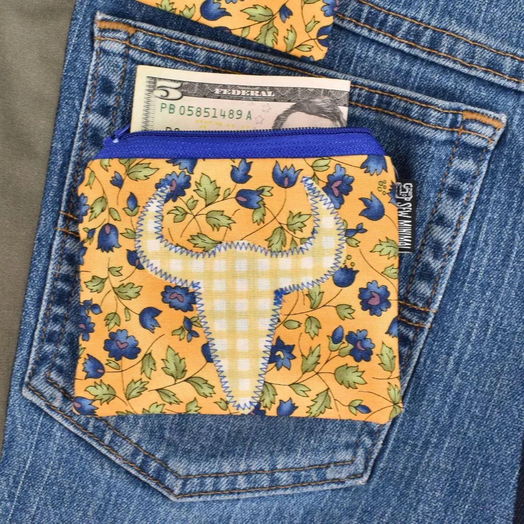 A yellow blue bell print pouch decorated with a tan check cow skull applique sits on a blue jean back pocket. A tube of chapstick is shown next to the pouch.