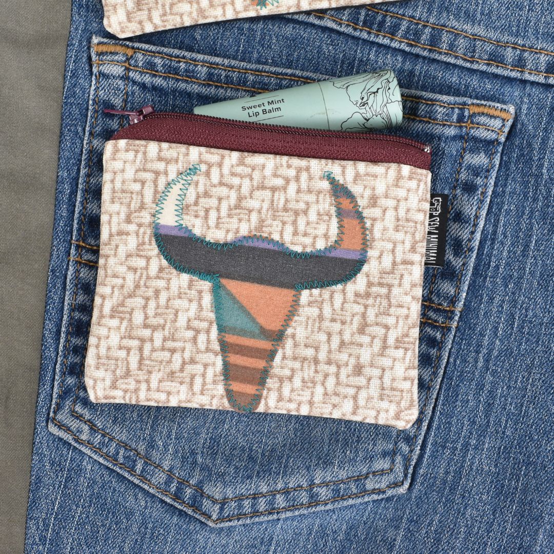 A beige pouch decorated with a southwest print cow skull applique sits on a blue jean back pocket. A tube of chapstick is shown next to the pouch.