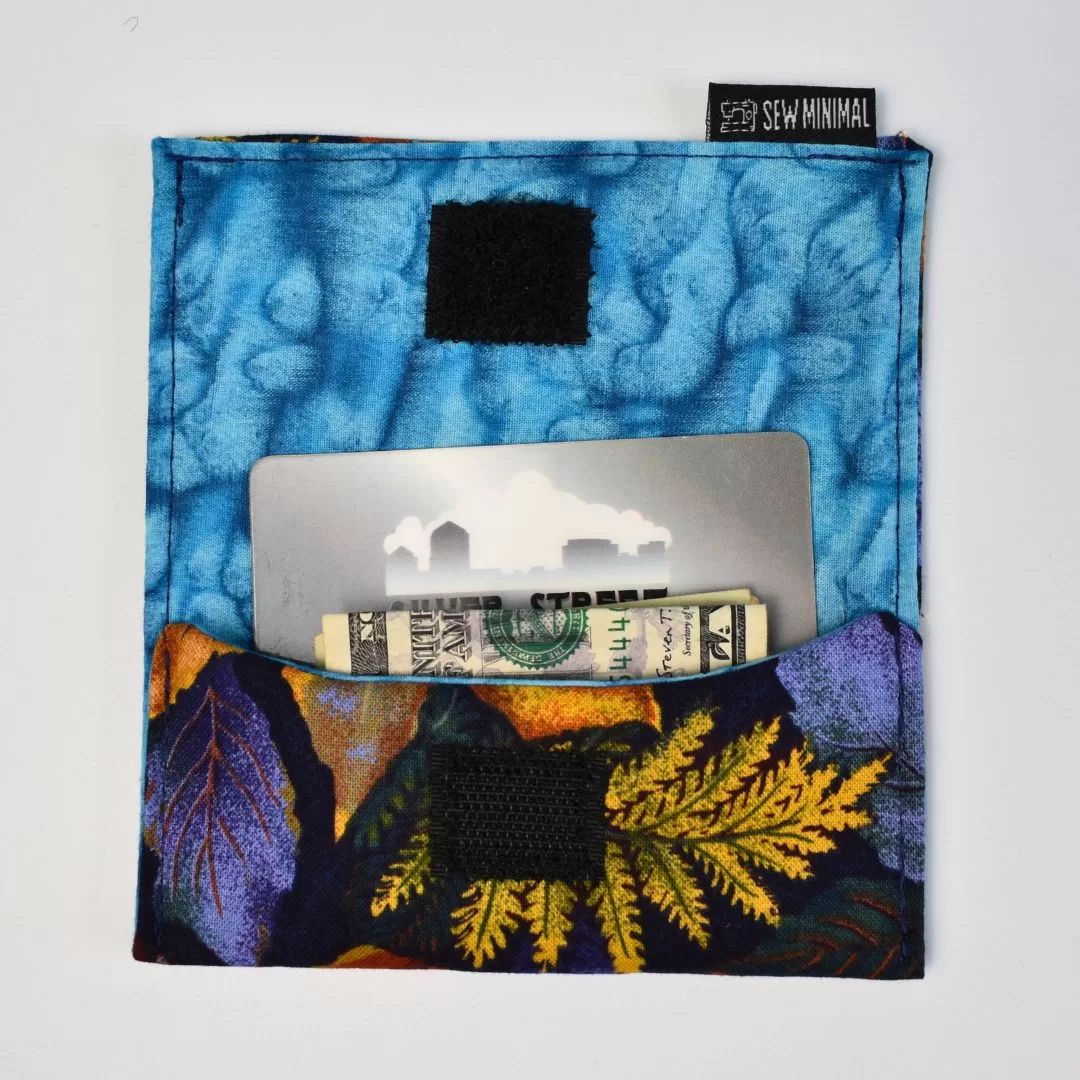 Blue and purple leaf print small cardholder wallet laying open with a dollar bill inside on a blank white background.