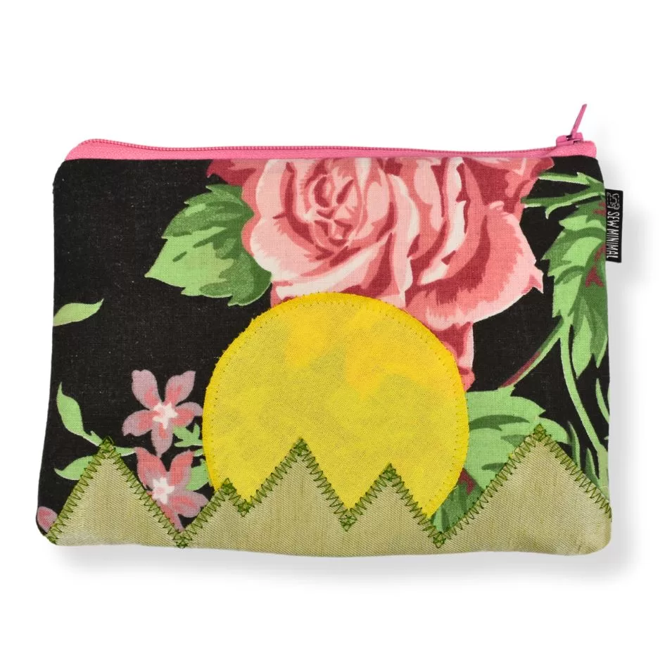 A small black and pink print fabric zipper pouch with a yellow sun and green mountain applique.