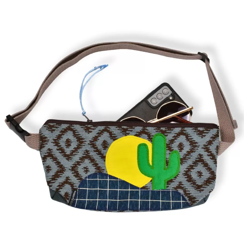 A woven convertible belt bag with a vibrant pattern and a cactus design is displayed with round sunglasses and a smartphone resting on top. The bag features a gray strap and a brown zipper.