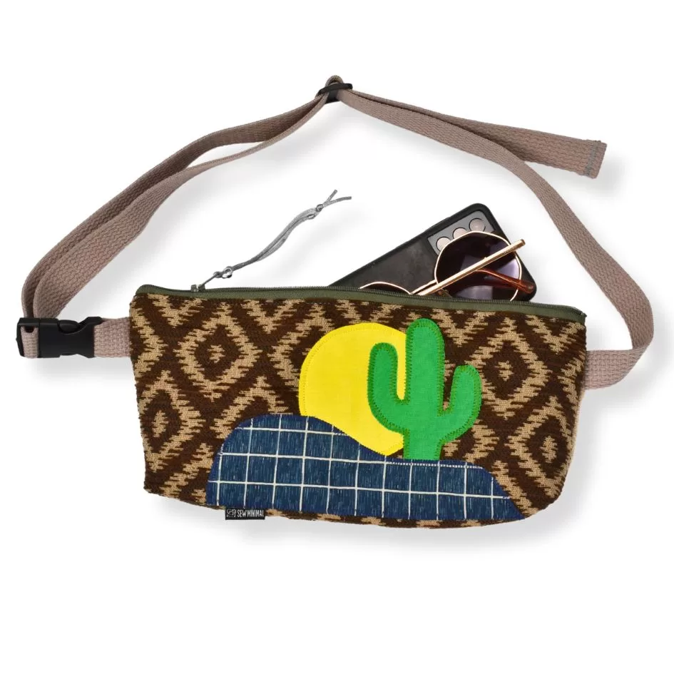 A woven convertible belt bag with a vibrant pattern and a cactus design is displayed with round sunglasses and a smartphone resting on top. The bag features a gray strap and an olive zipper.