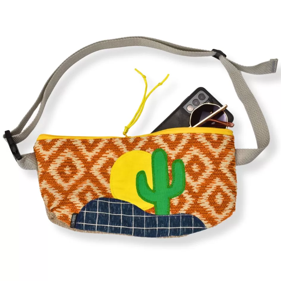 A woven convertible belt bag with a vibrant pattern and a cactus design is displayed with round sunglasses and a smartphone resting on top. The bag features a gray strap and a yellow zipper.