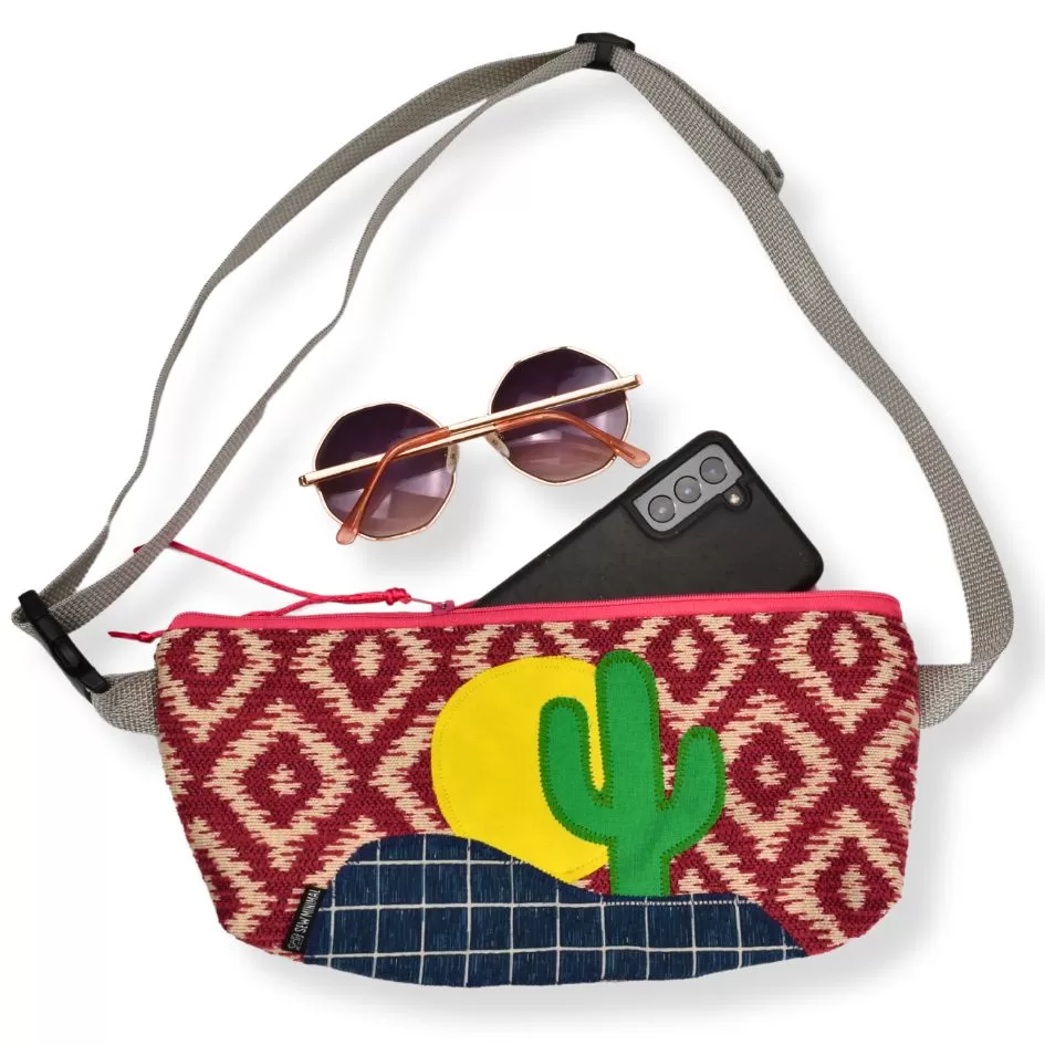 A woven convertible belt bag with a vibrant pattern and a cactus design is displayed with round sunglasses and a smartphone resting on top. The bag features a gray strap and a pink zipper.