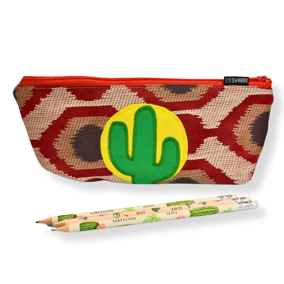 A Cactus Pencil Holder Pouch with a red and gold snakeskin pattern, featuring a green cactus in a yellow circle on the front. Two pencils with a succulent pattern are placed in front of the pencil case.