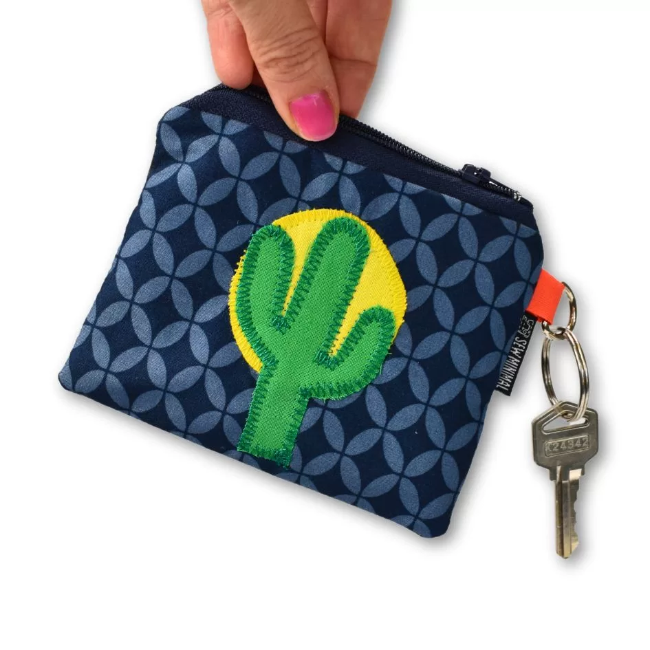 A hand holding a small key ring pouch with a blue geometric pattern and a green cactus patch stitched on a yellow background. The pouch has a zipper and an attached key with a labeled keychain.