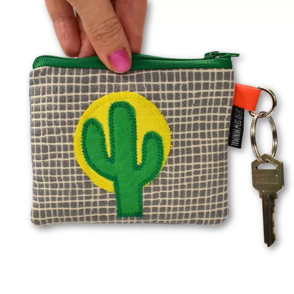 A hand holding a small key ring pouch with a gray square pattern and a green cactus patch stitched on a yellow background. The pouch has a zipper and an attached key with a labeled keychain.