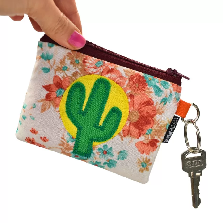 A hand holding a small key ring pouch with a pink floral pattern and a green cactus patch stitched on a yellow background. The pouch has a zipper and an attached key with a labeled keychain.