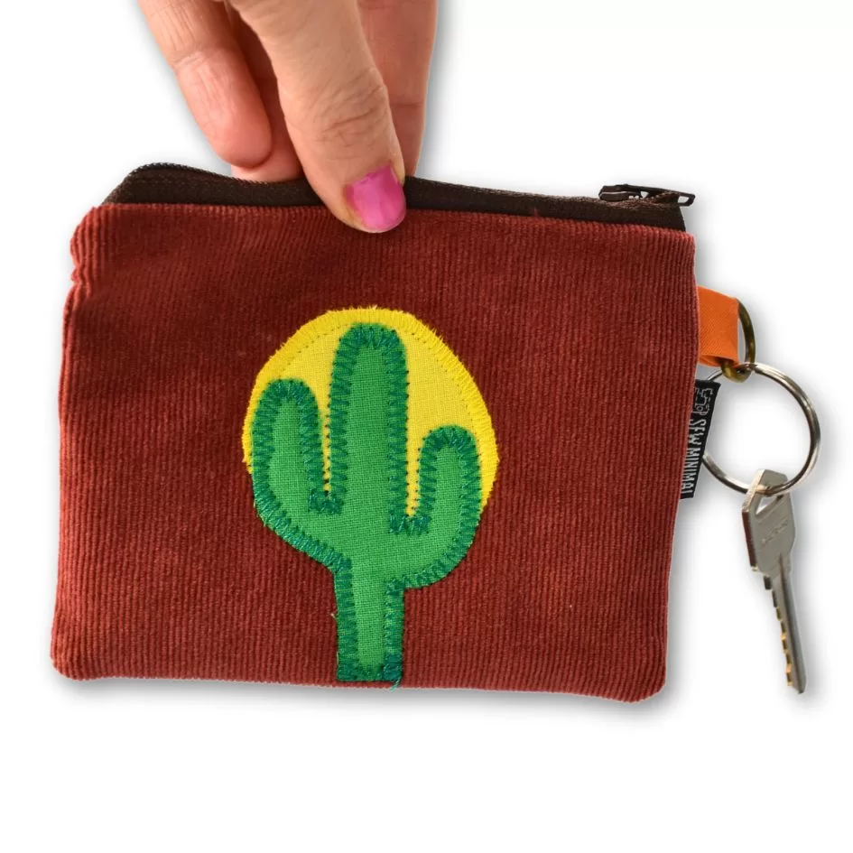 A hand holding a small key ring pouch with a red corduroy pattern and a green cactus patch stitched on a yellow background. The pouch has a zipper and an attached key with a labeled keychain.