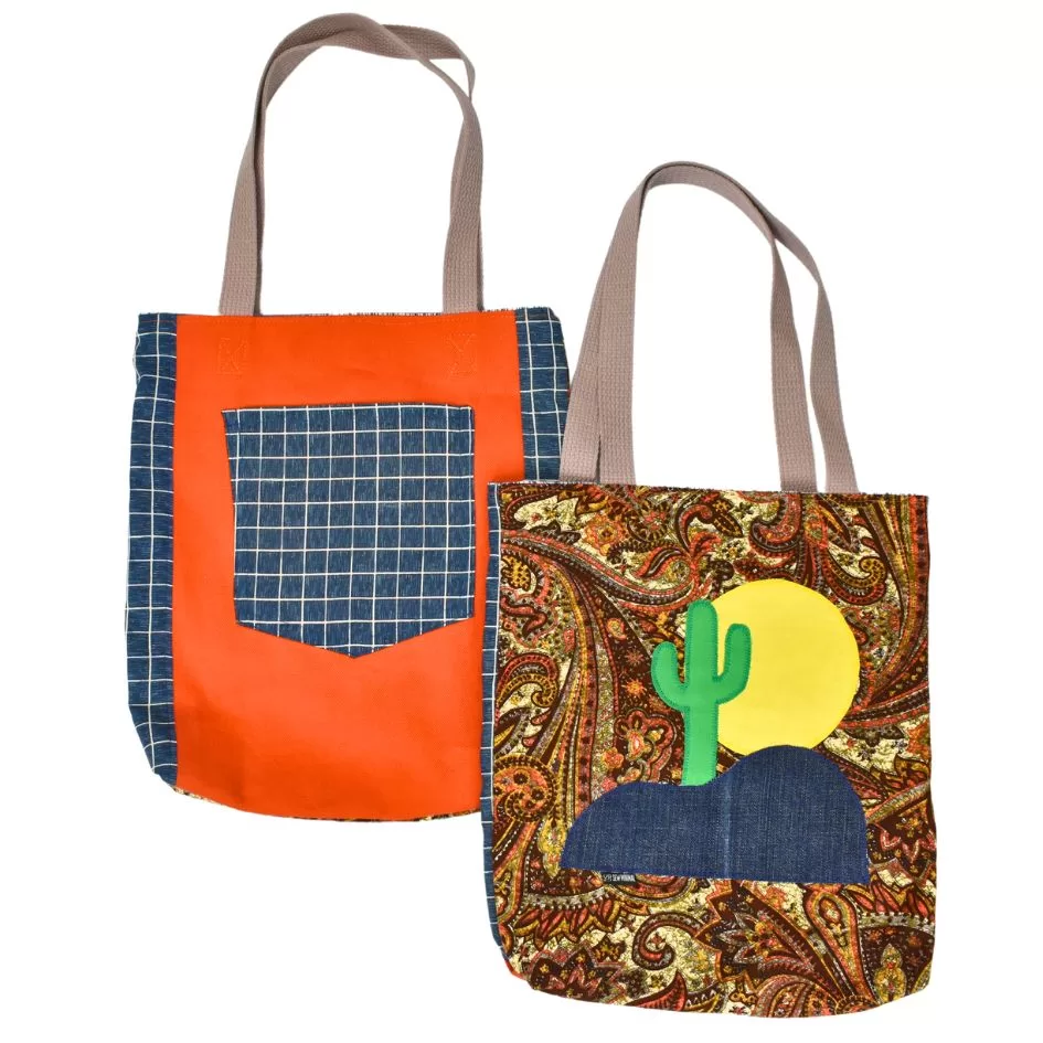 The front and back side of the cactus tote bag with pockets. The left bag shows the back side of the tote which features a bold orange canvas with a blue checkered pocket, while the right bag showcases the front of the tote with paisley pattern and an appliqué of a cactus, sun, and denim hill. The bags have gray handles.