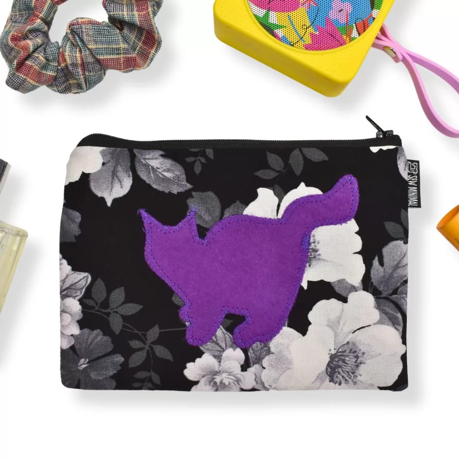 The image shows a black zippered pouch with a floral pattern in shades of white and gray, featuring a bold purple silhouette of a cat in the center. Surrounding the pouch are various small items, including a plaid scrunchie, a yellow box with colorful contents, and other accessories like perfume, suggesting a stylish and organized setup.