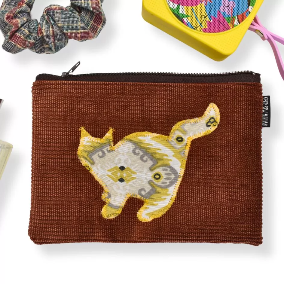 The image shows a brown zippered pouch with a textured fabric, featuring a patterned yellow and white silhouette of a cat in the center. Similar to the previous image, it is surrounded by small items such as a plaid scrunchie, a yellow box with colorful contents, and other accessories, like perfume, creating a stylish and coordinated display.