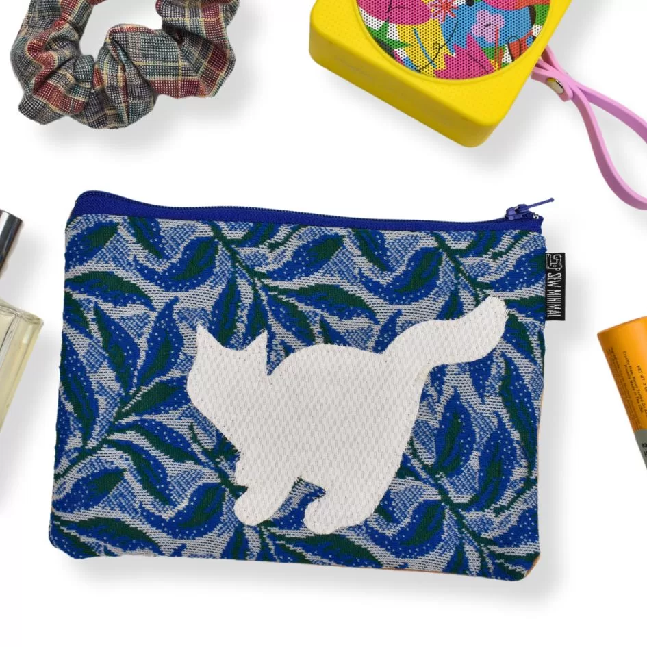 This image shows a pouch with a blue and green leaf pattern, featuring a white silhouette of a cat in the center. Surrounding the pouch are various items, including a plaid scrunchie, a colorful speaker, perfume, and a tube of orange skincare product. The layout suggests a flat-lay style, possibly showcasing everyday accessories or travel essentials.