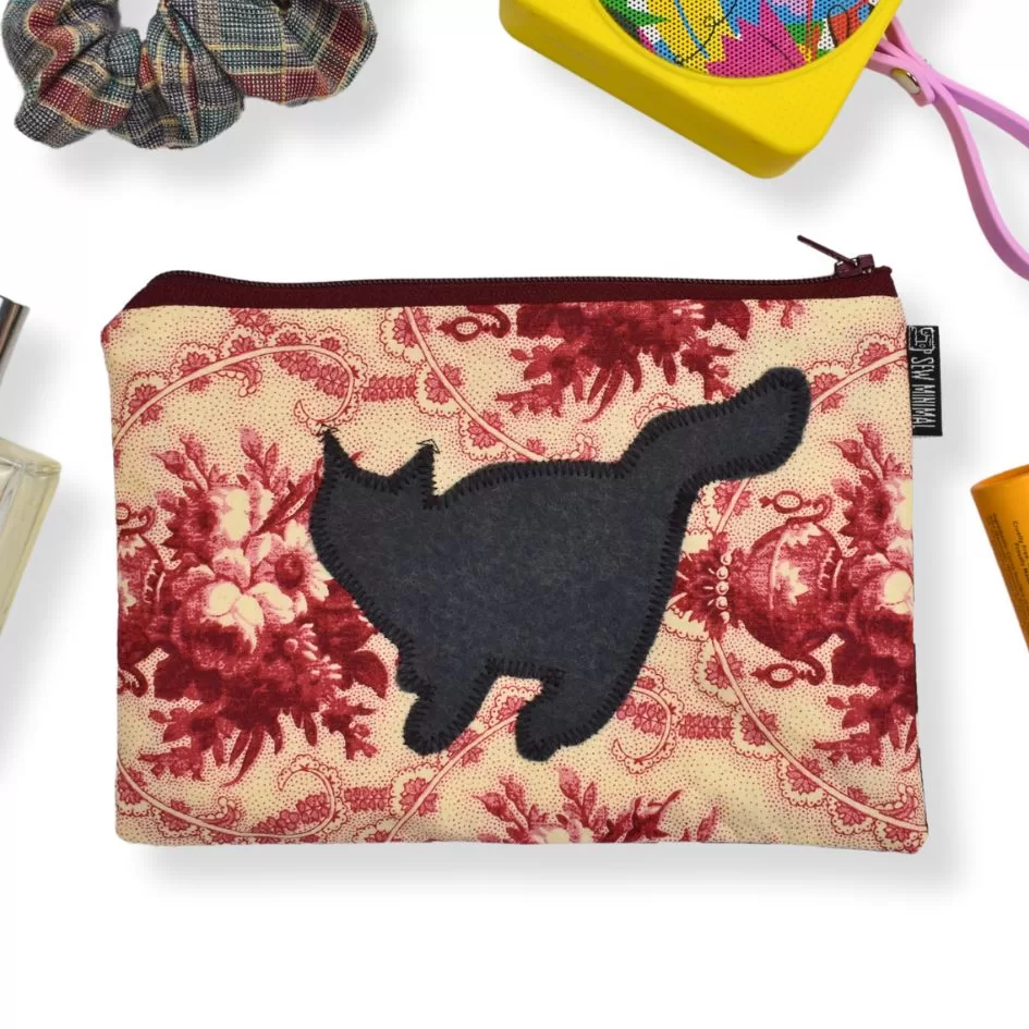 This image shows a pouch with a cream background and a maroon floral pattern, featuring a black silhouette of a cat in the center. Similar to the previous image, the pouch is surrounded by items such as a plaid scrunchie, a colorful speaker, perfume, and an orange skincare product. The arrangement is in a flat-lay style, emphasizing everyday accessories or travel essentials.