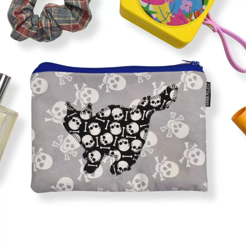 This image shows a pouch with a grey background featuring white skull and crossbones patterns. In the center, there is a black silhouette of a cat, filled with white skulls and bones. The pouch is surrounded by a plaid scrunchie, a colorful speaker, perfume, and an orange skincare product, arranged in a flat-lay style, emphasizing a quirky or edgy aesthetic with everyday accessories.