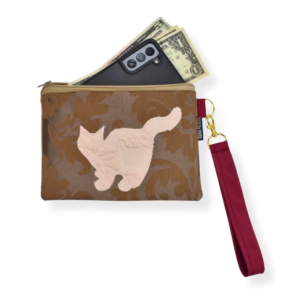 The image shows a small zippered pouch with a floral pattern in brown fabric, featuring an applique of a cat on the front. The pouch contains a smartphone and U.S. dollar bills partially sticking out. It has a maroon wrist strap attached by a gold-tone clip.