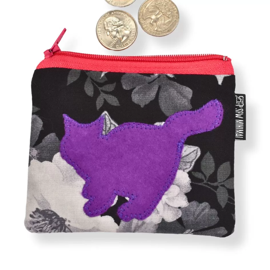 The image shows a small black and white floral fabric pouch with a bright red zipper. A purple fabric patch in the shape of a cat is sewn onto the front of the pouch. Three U.S. quarters are positioned near the top of the pouch, suggesting the pouch is being used for storing coins or small items. There is a fabric tag on the side that reads "SEW MINIMAL."