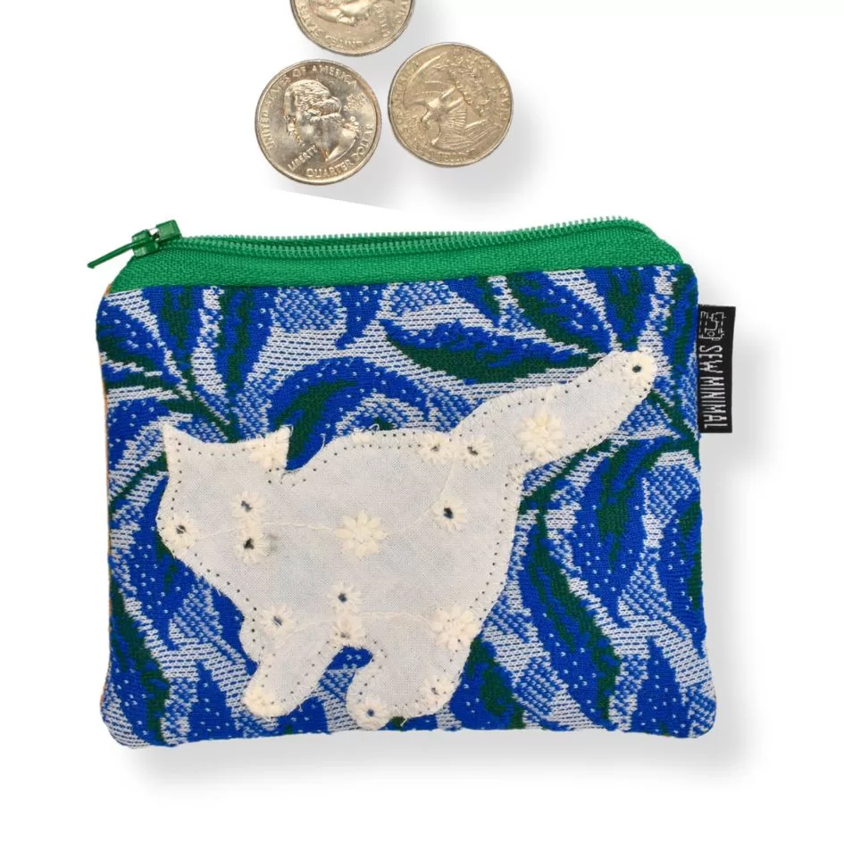 This image shows a small, decorative coin pouch with a green zipper. The fabric features a white cat applique stitched onto a blue and green leafy background. A few U.S. quarters are placed above the pouch, indicating its potential use for storing small items like coins.