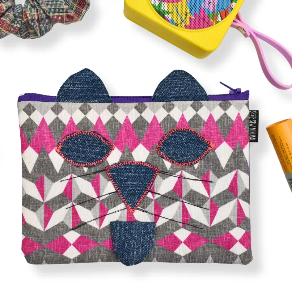 This image features a small fabric pouch designed with a geometric pattern in pink, white, and gray, resembling a cat's face. The pouch has denim details for the cat's ears, nose, and mouth, adding a playful touch to the design. Surrounding the pouch are colorful items including a plaid scrunchie, a yellow container with a rainbow pattern, and part of an orange cylindrical object.