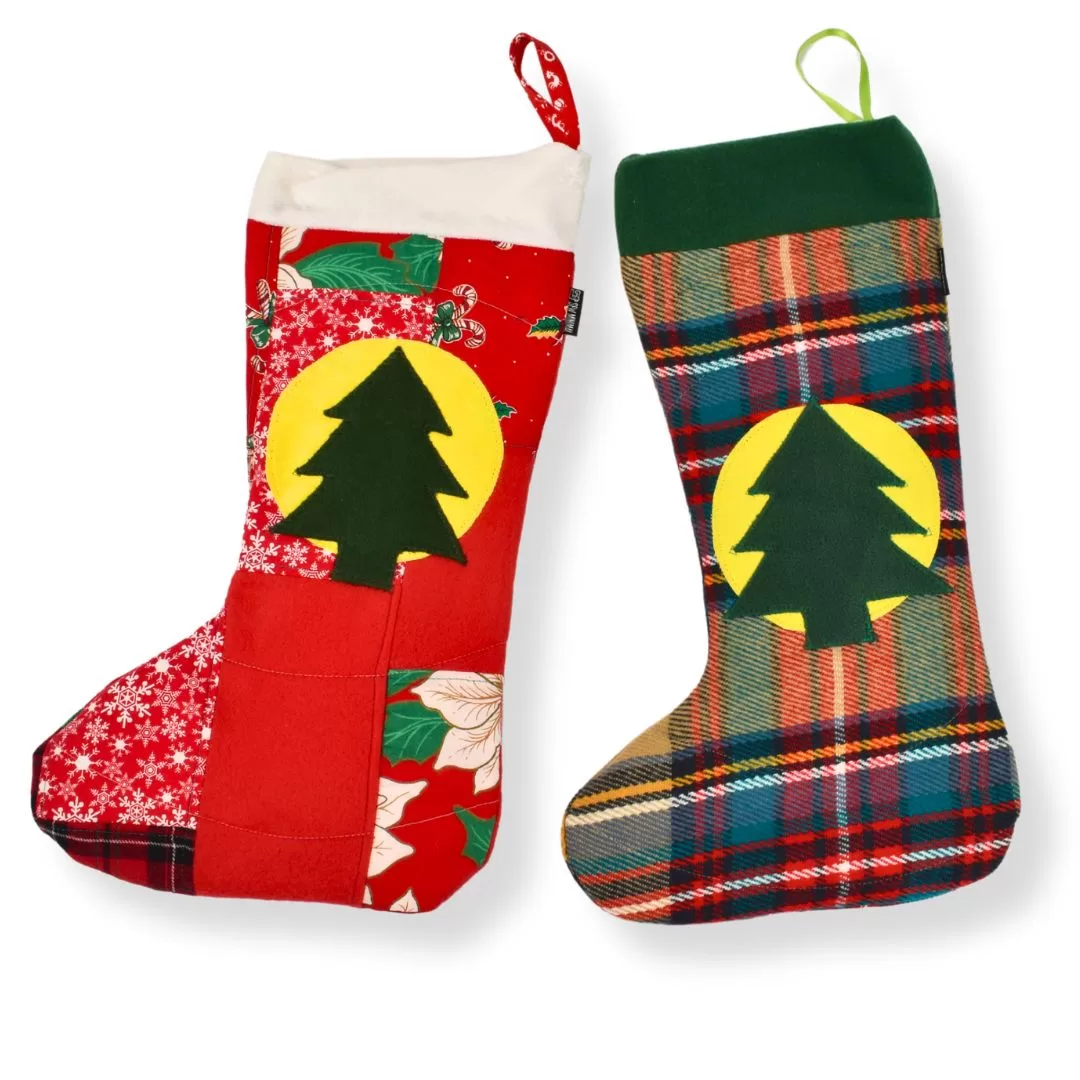 Two handmade Christmas stockings crafted from upcycled textiles. The left stocking features a red patchwork design with festive floral patterns and a green tree applique on a yellow circular background, paired with a red hanging loop. The right stocking showcases a plaid design in green, red, and yellow tones with a matching tree applique and a green hanging loop.